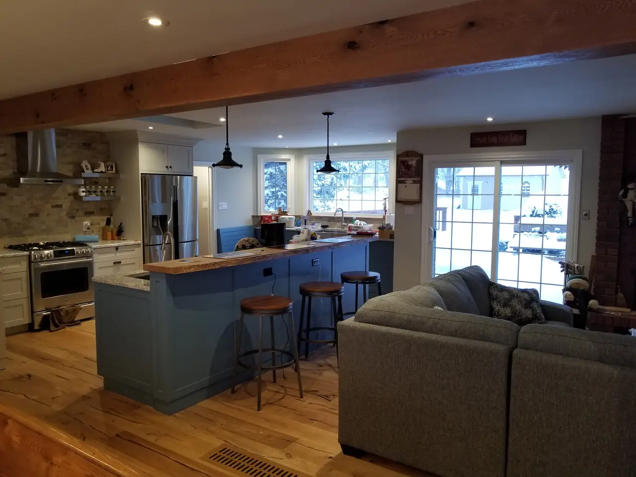 Evening view of a renovated open-concept main floor: kitchen island with stools, exposed timber beam, and patio doors to a snowy yard.